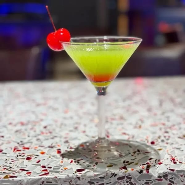 A vibrant green and red cocktail in a martini glass, garnished with a cherry, on a speckled table.