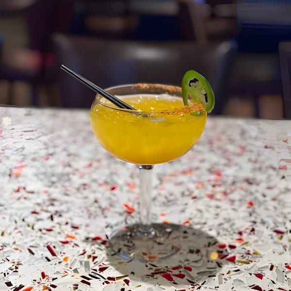 A yellow cocktail with a chili-salted rim and a jalapeño slice sits on a speckled white table.