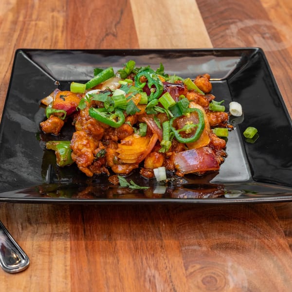 Masthi Bar & Grill's Indian chili chicken, garnished with green chilies, onions, and cilantro on a black plate.