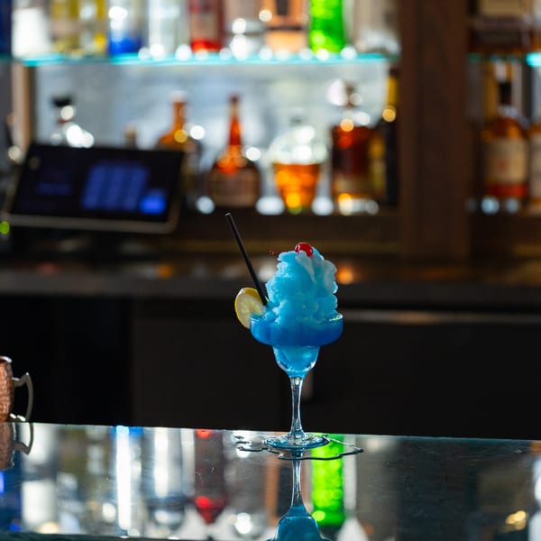 A vibrant blue frozen cocktail with a cherry and lemon slice sits on a reflective bar.
