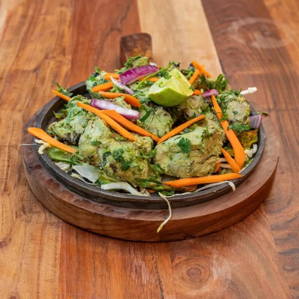 Indian Hariyali Chicken Tikka, green marinated chicken pieces with carrots, onions, and lime on a sizzling platter.