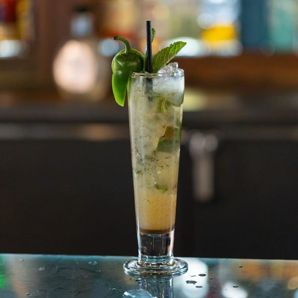 A tall glass of iced mojito garnished with a green pepper and mint leaves on a bar.