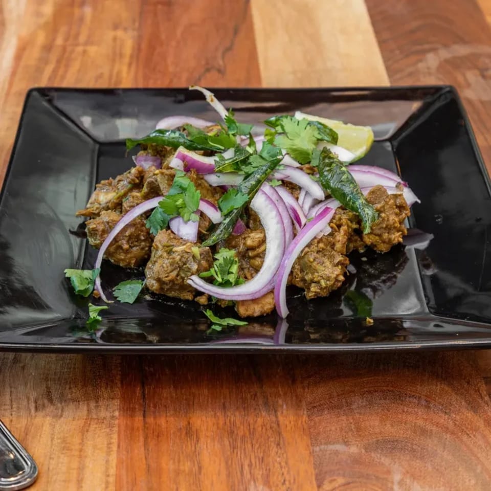 Indian chicken fry, garnished with red onions, cilantro, and a lime wedge on a black plate.