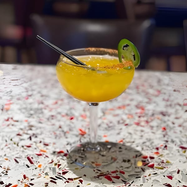 A vibrant yellow cocktail with a chili-salt rim, black straw, and jalapeño slice on a speckled white table.