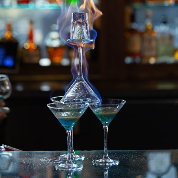 Flaming blue cocktails stacked in martini glasses on a bar counter with bottles in the background.